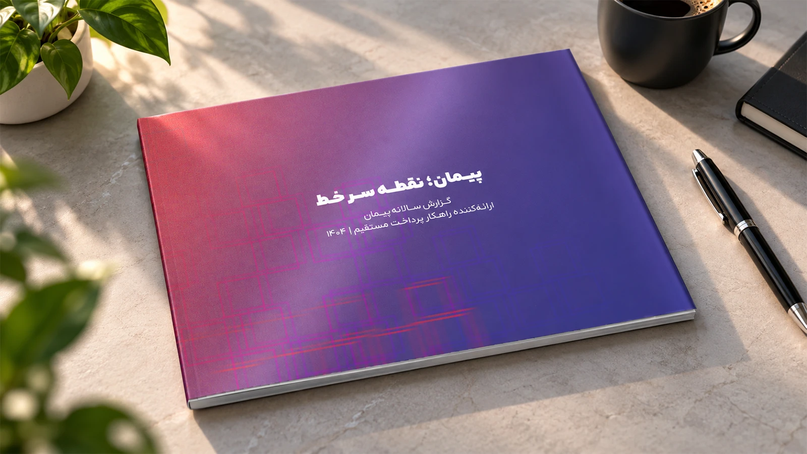 جلد گزارش سالانه ۱۴۰۴ پیمان با طراحی بنفش و صورتی روی میز روشن، در کنار فنجان قهوه، خودکار مشکی، دفترچه و گیاه سبز
